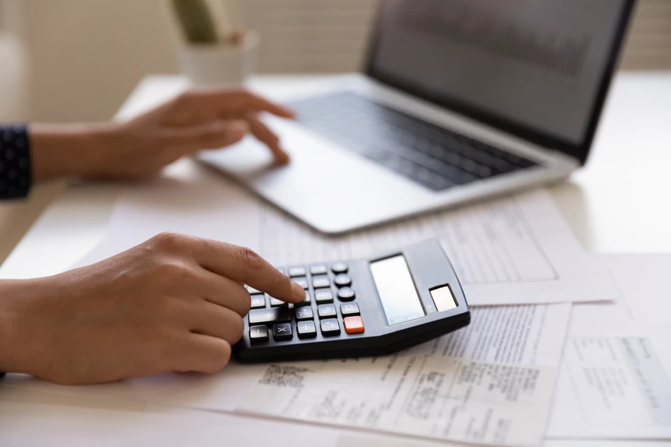 Personne travaillant dans un bureau utilisant une calculatrice pour analyser des documents financiers, avec un ordinateur portable ouvert sur un bureau blanc