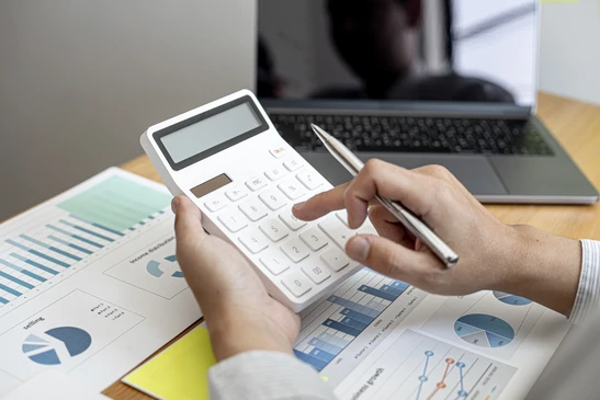 Personne utilisant une calculatrice blanche dans un bureau, tenant un stylo, analysant des rapports financiers avec graphiques, tableaux et diagrammes circulaires étalés sur un bureau en bois